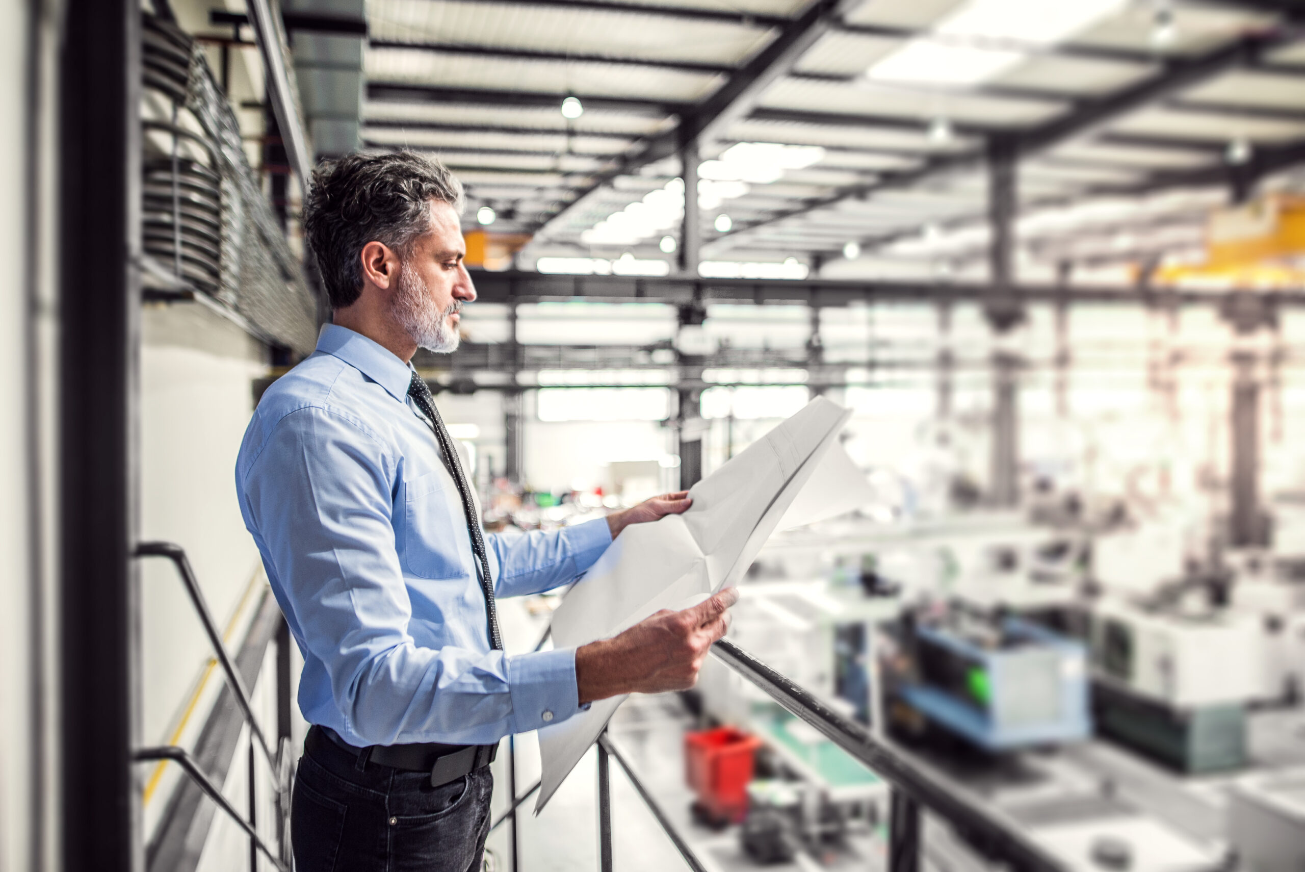 Portrait of project manager reading blueprint modern industrial factory. Manufacturing facility with robotics, robotic arms and automation. Storing products and materials in warehouse.